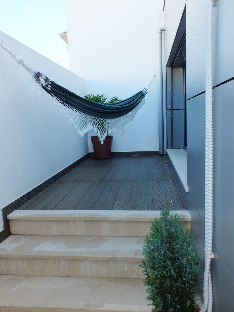 Small private patio with a black-and-white hammock strung between two walls, a potted palm in the corner, and steps leading up to a wood-look deck beside a sliding door.