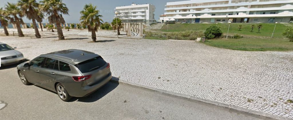 Cobblestone parking area near the seafront in Buarcos with palm trees, a parked car, and access to the beach walkway